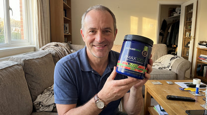 A man in a blue polo shirt holds up a tub of Pofassi Max Pmax1000 protein powder in a living room setting.